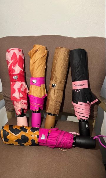 Surtido de paraguas plegados en rosa brillante, naranja, negro y gris junto a una cesta tejida y hojas verdes.
