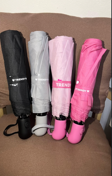 Cuatro paraguas compactos plegados en negro, gris, rosa y rosa fucsia, apoyados verticalmente contra una pared.
