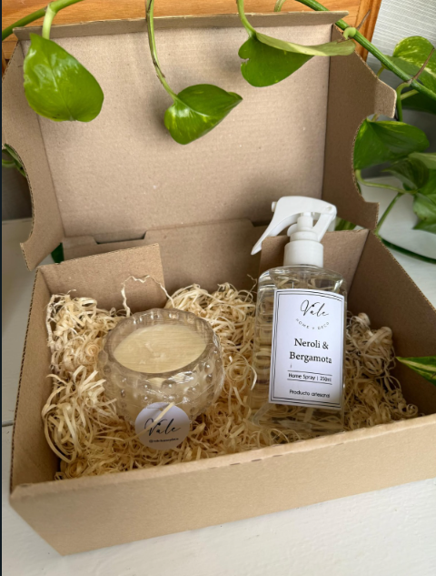 Caja de regalo con una vela y un pulverizador envueltos en paja, rodeados de hojas verdes de plantas trepadoras.