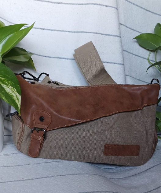 Bolso de hombro de piel marrón y gris con correa sobre una superficie a rayas, enmarcado por hojas verdes.