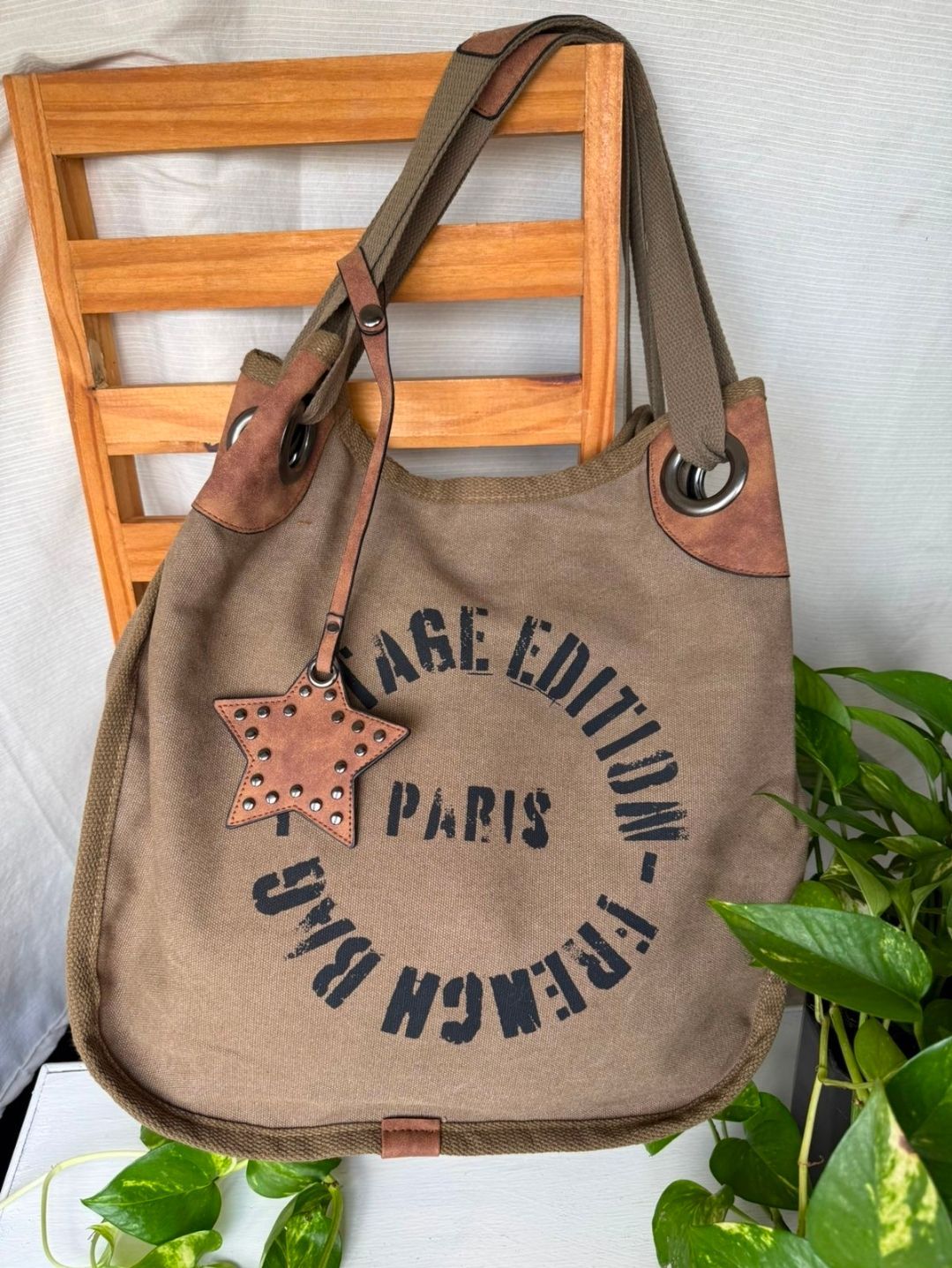 Bolso de lona marrón con texto negro colgado en una silla de madera junto a hojas verdes.