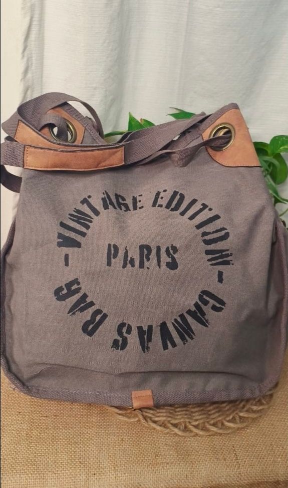 Bolso de lona marrón con texto negro colgado en una silla de madera junto a hojas verdes.
