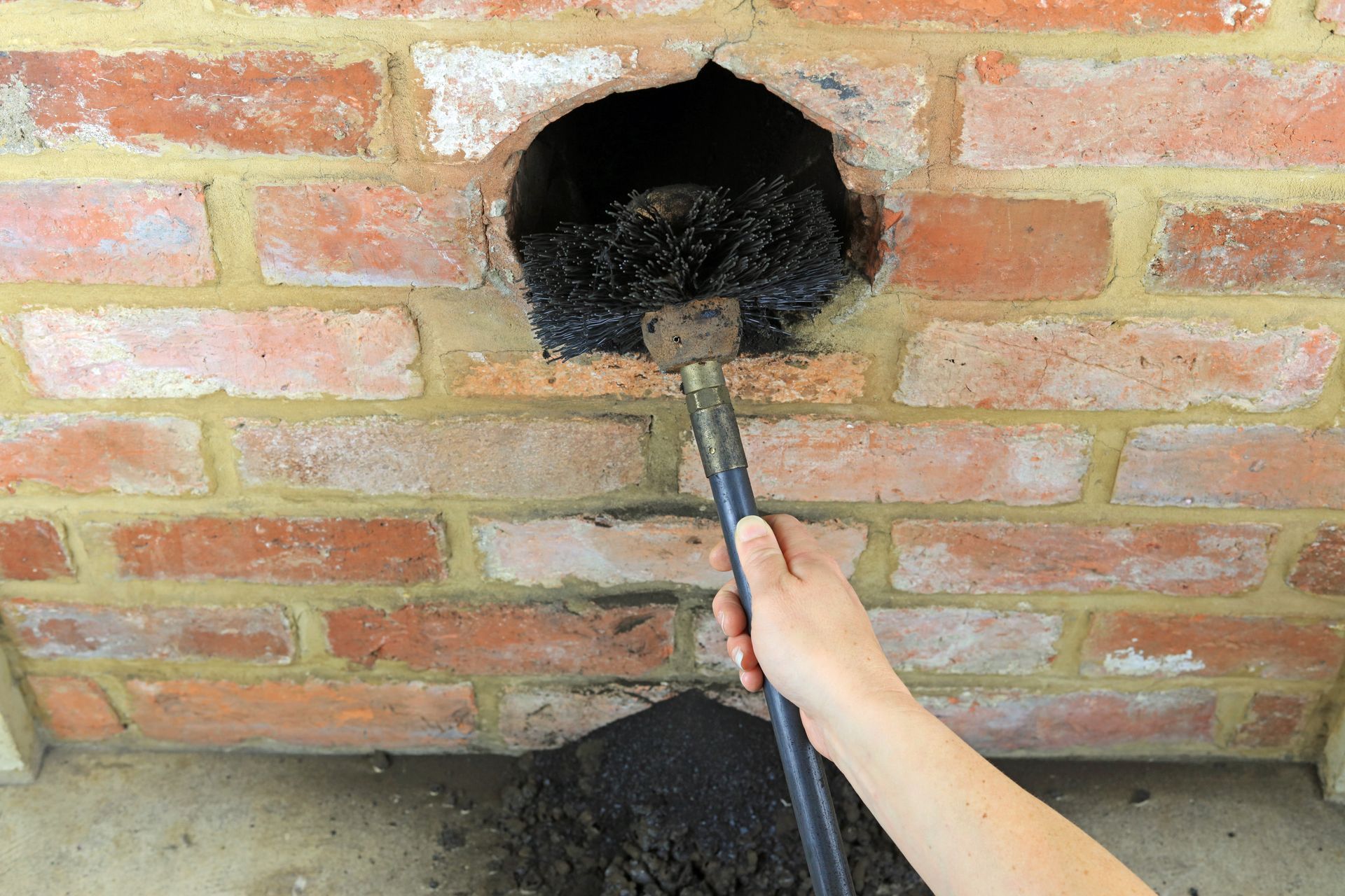 Person cleaning a brick wall vent hole with a long brush and hose Person cleaning a brick wall vent with a brush and hose leading into a round hole