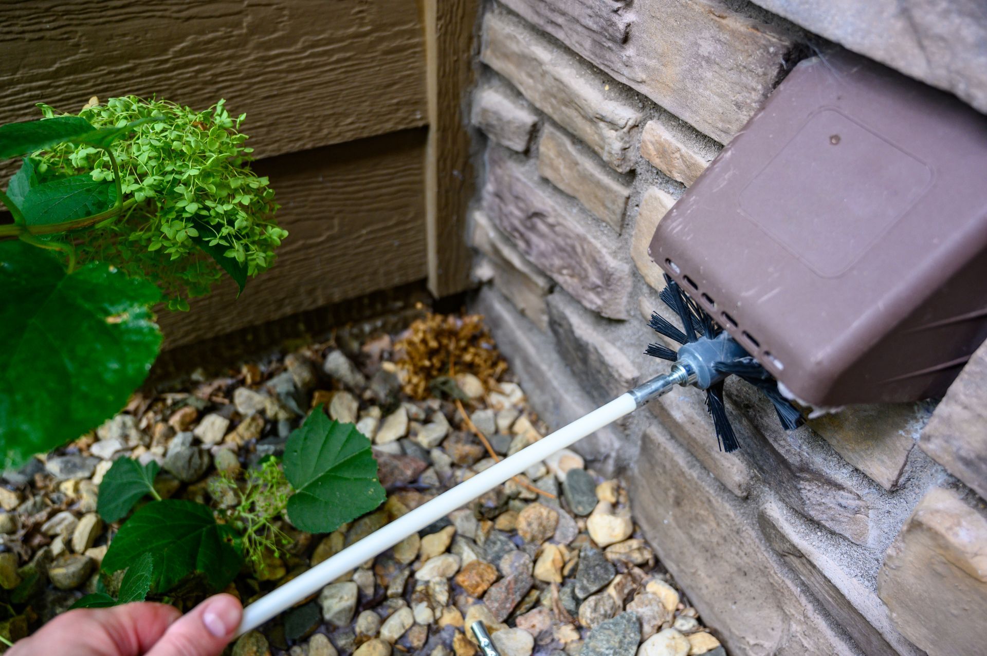 Hand holding a garden hose spraying water near a home's exterior wall and foundation rocks Hand holding a garden hose spraying water near a home's exterior wall and foundation rocks
