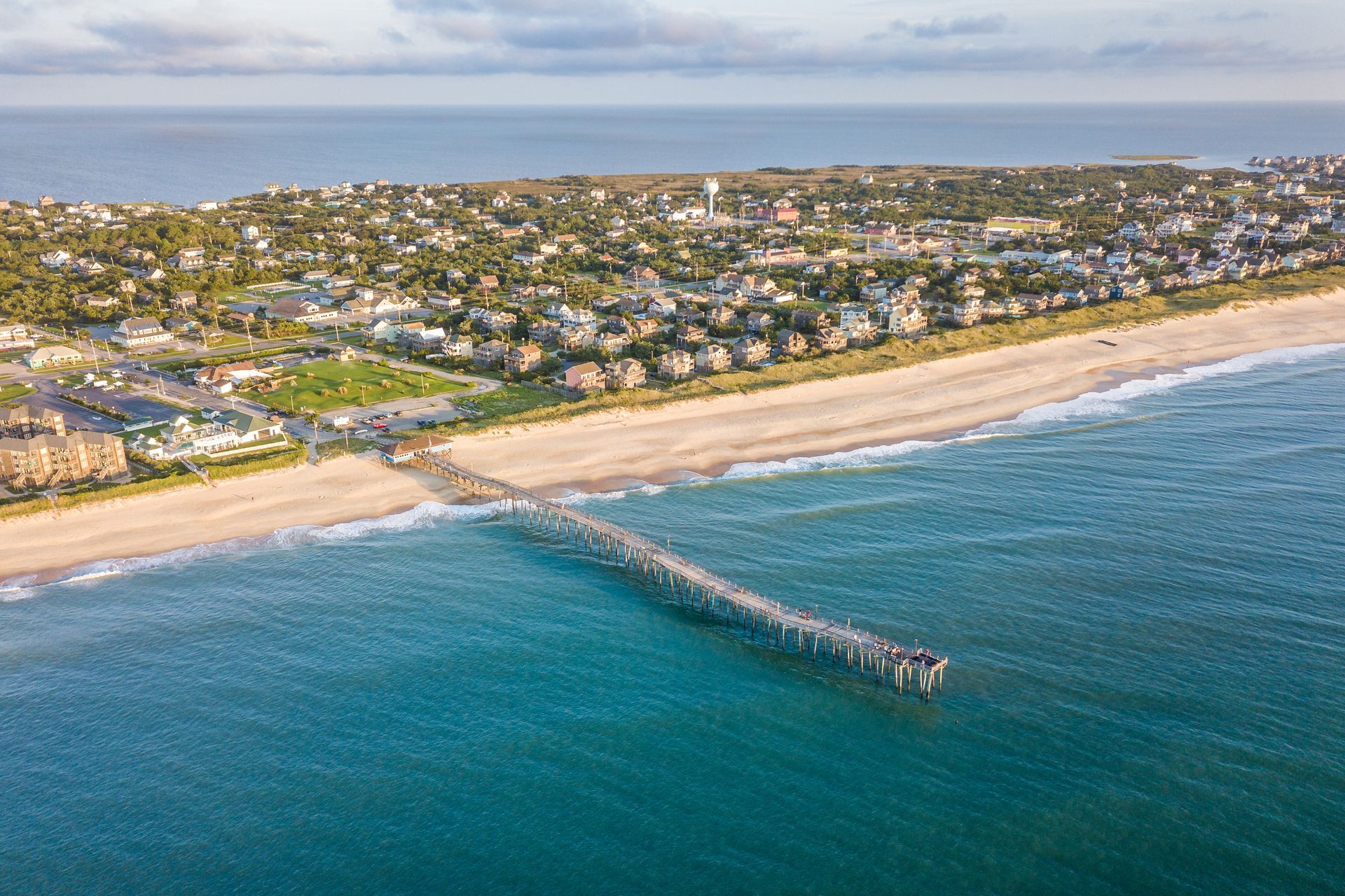 Aerial view of a coastal town with a long pier extending into blue ocean beside sandy beaches Aerial view of a coastal town with a sandy beach and a long pier extending into blue water