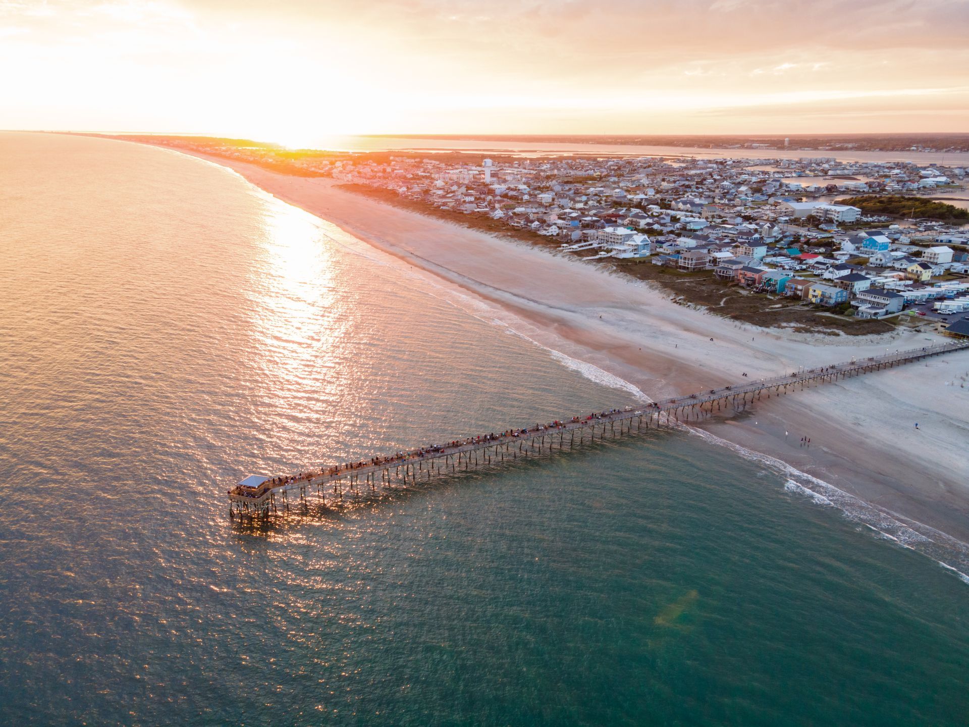 Aerial view of a beach at sunset with waves, a long jetty, and coastal buildings along the shore Aerial view of a beach at sunset with waves, a long pier, and a coastal town along the shore