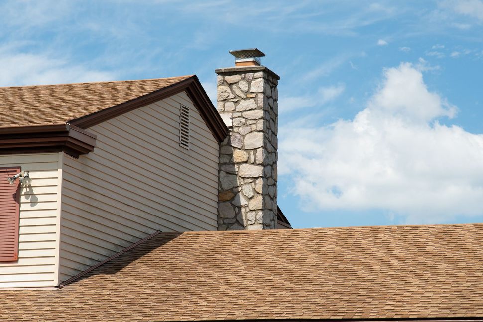 Stone chimney on a tan shingle-roofed house against a blue sky with clouds