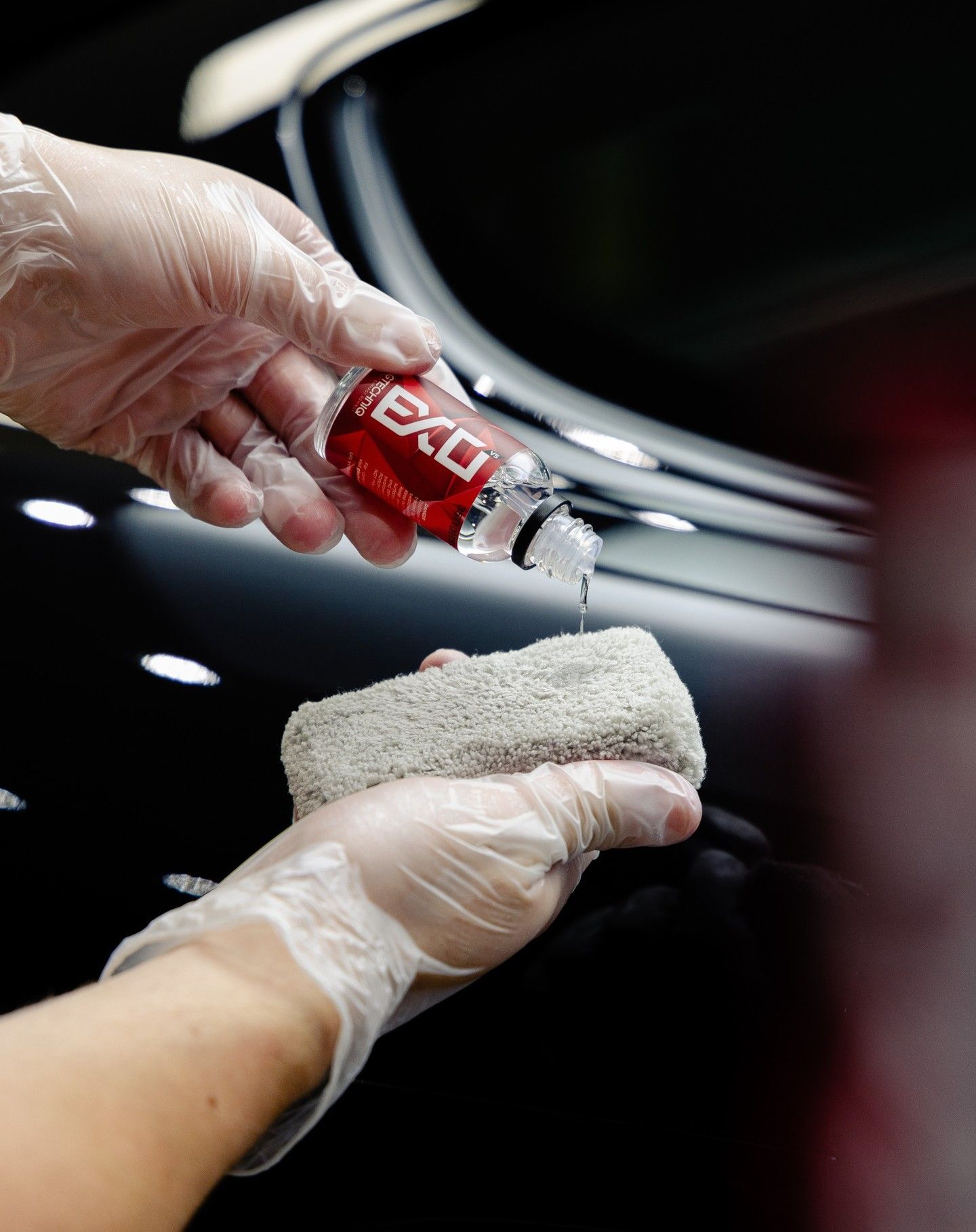 Gloved hands pouring red car wash liquid onto a sponge over a black car surface
