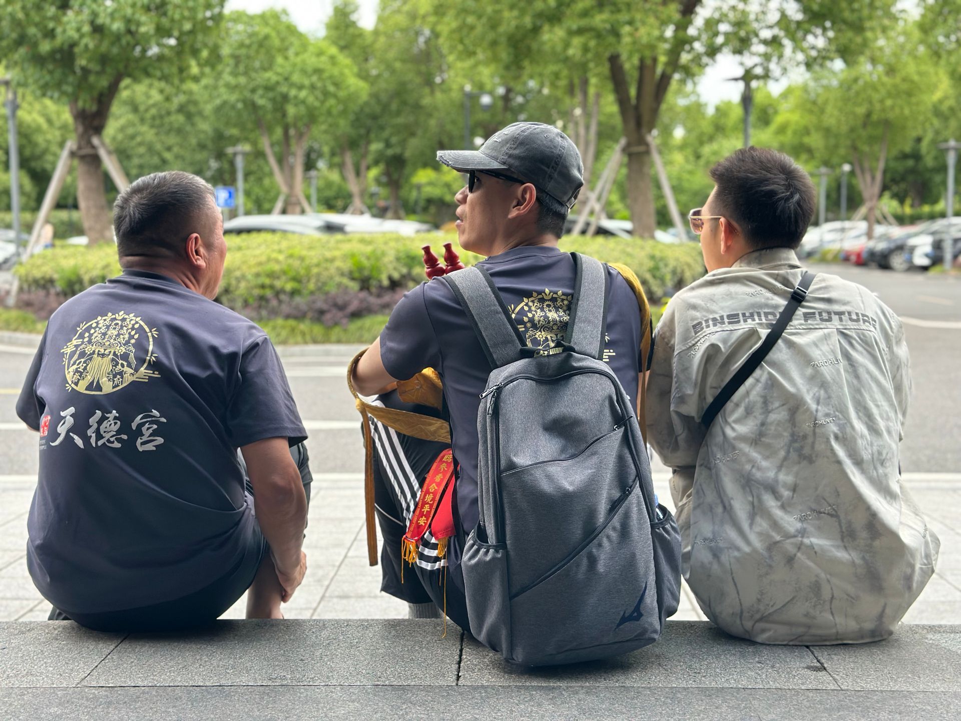三個人坐在公園的長椅上，背對著鏡頭，背景是背包和樹木。