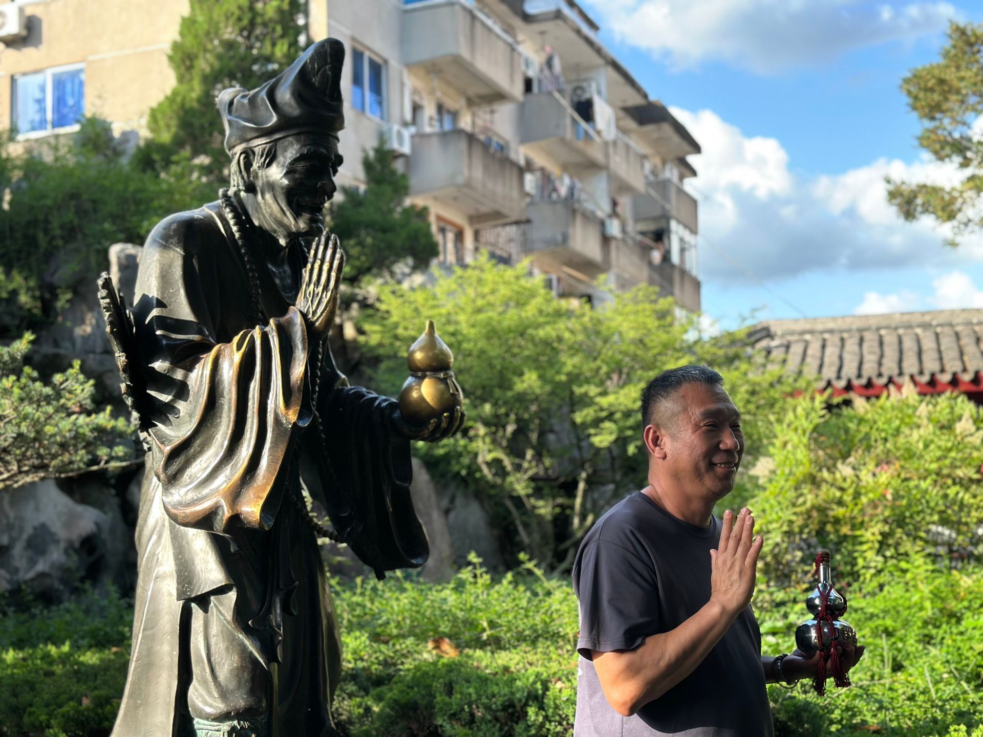 男子在鬱鬱蔥蔥的花園中，在一尊黑色雕像旁祈禱，花園裡有建築物和藍天。