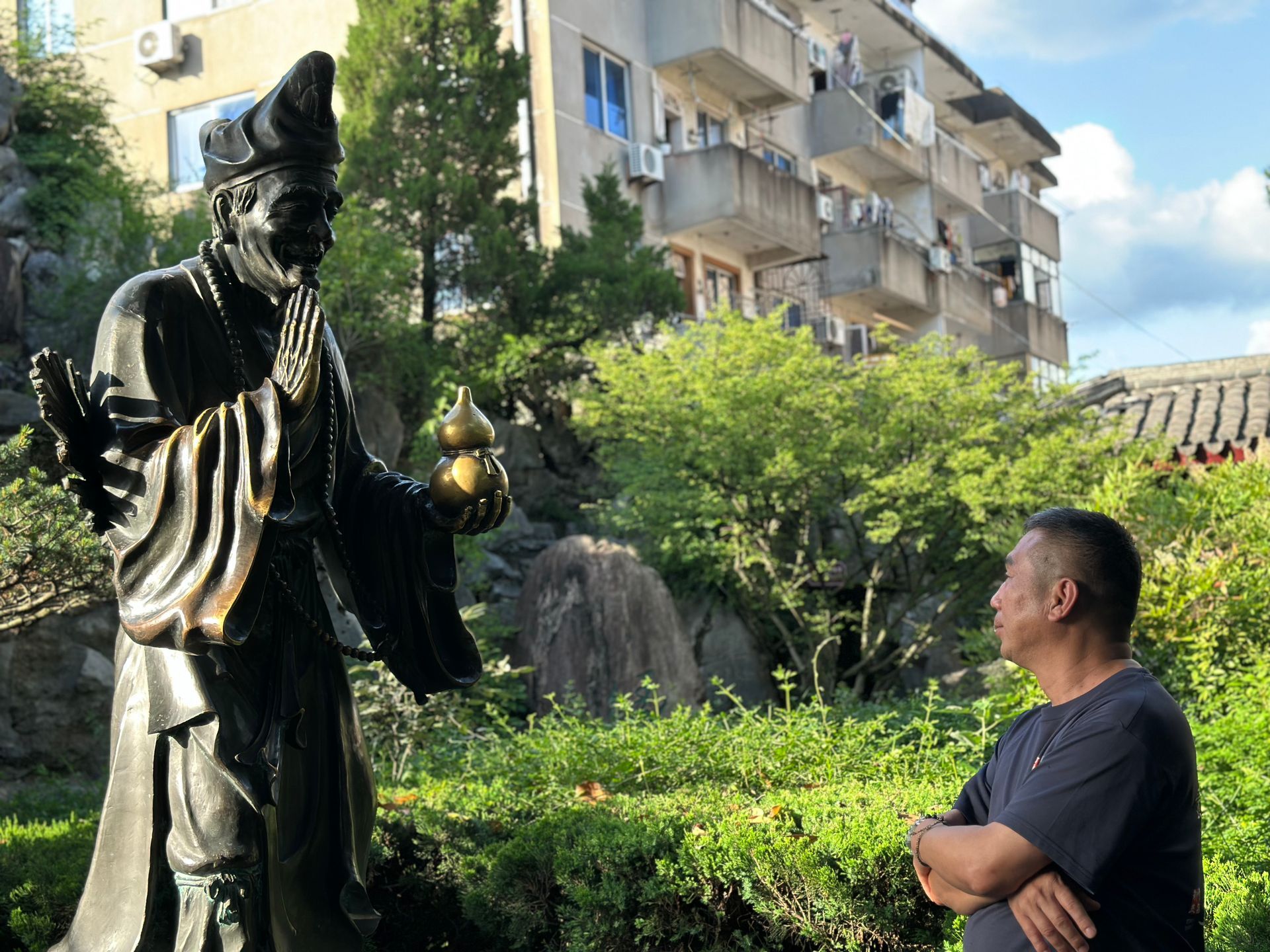 一個男人站在花園裡一座黑色雕像旁，身後是公寓大樓和樹木。