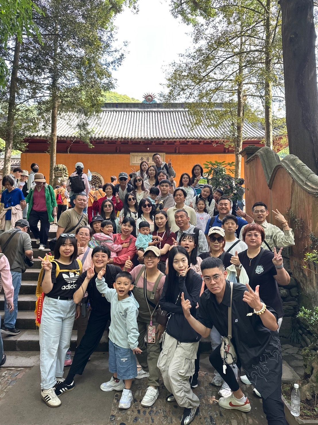 一大群人在戶外，在一棟紅色建築和樹木前擺姿勢拍照，許多人面帶微笑，向鏡頭揮手。