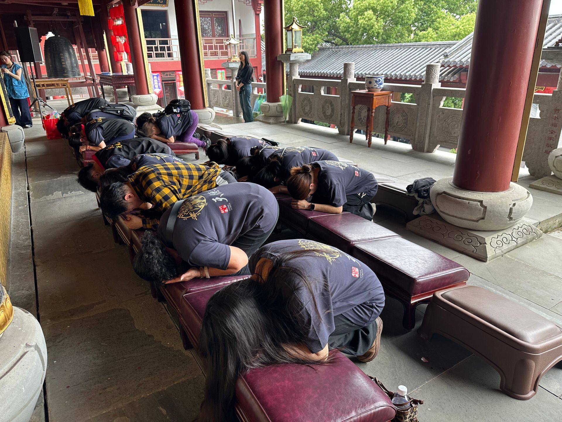 人們在寺廟庭院的長凳上深深鞠躬，背景是紅色的柱子和石板路。