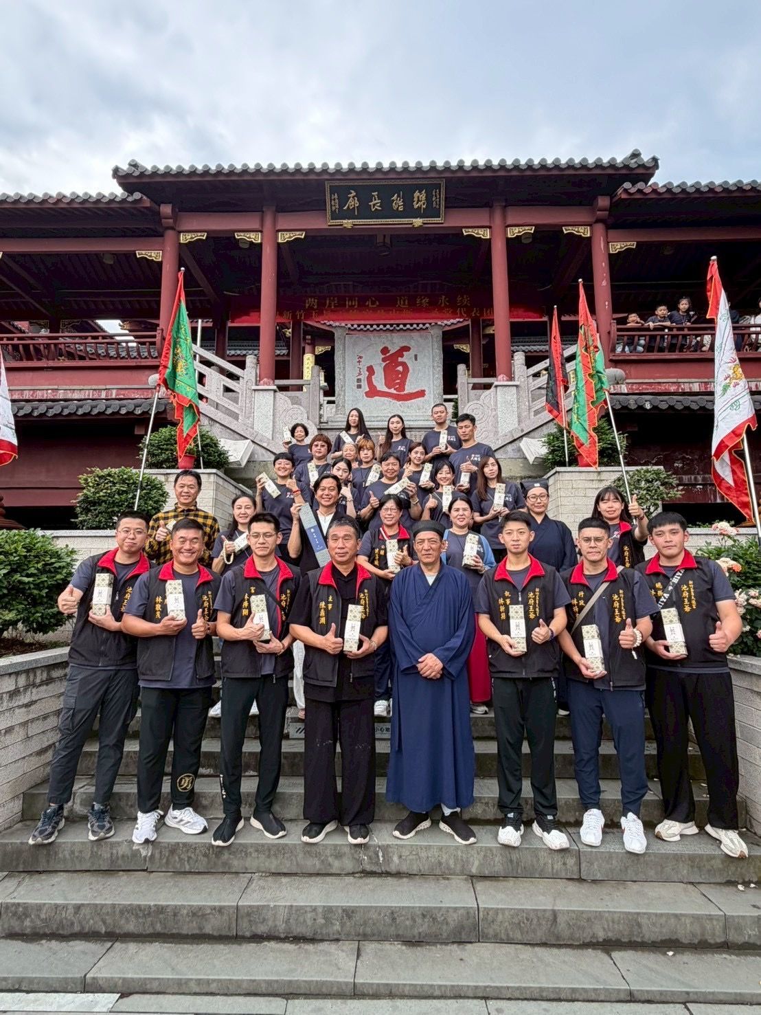一群人手持旗幟，在寺廟階梯上合影，背景是一座紅金相間的中國式建築。
