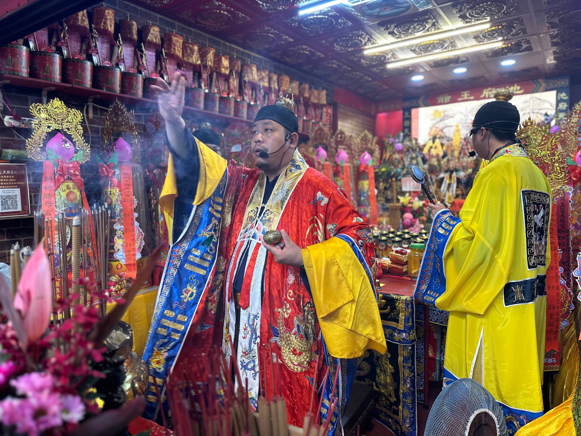 鮮花簇擁的神社裡，人們身著色彩鮮豔的僧袍，其中一人手持黃色扇子，沐浴在明亮的燈光下。