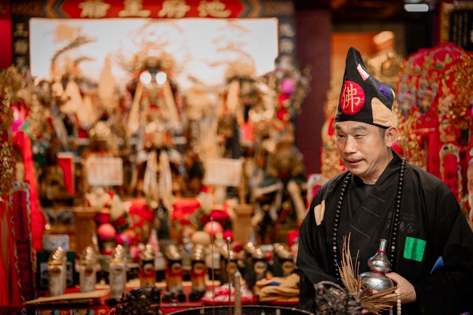 身穿黑袍、頭戴黑帽的男子站在擺滿雕像和祭品的紅色祭壇前。