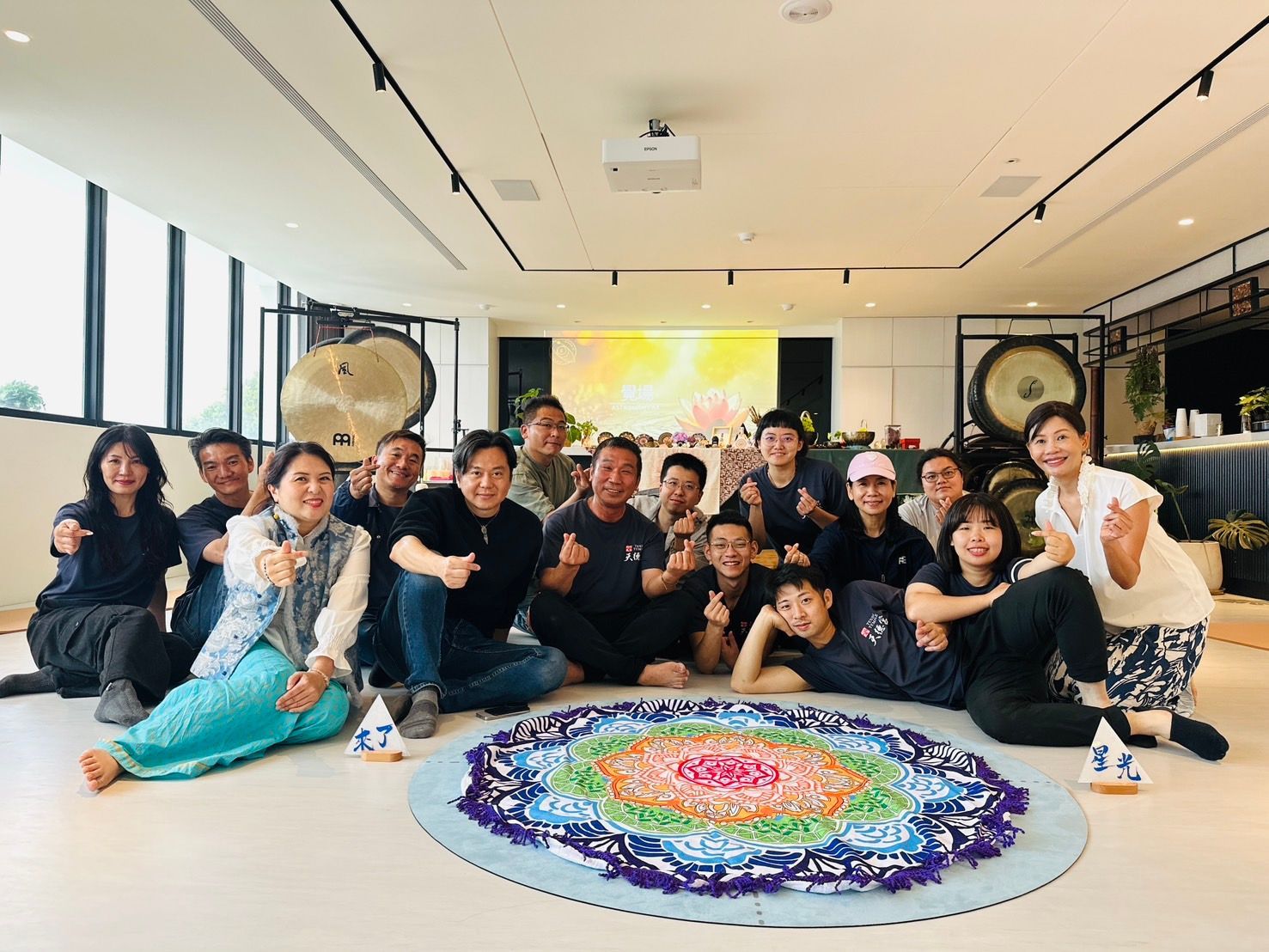 一群人在明亮的畫廊裡，圍繞著一幅色彩繽紛的圓形地面藝術品擺姿勢，面帶微笑，並肩而坐。