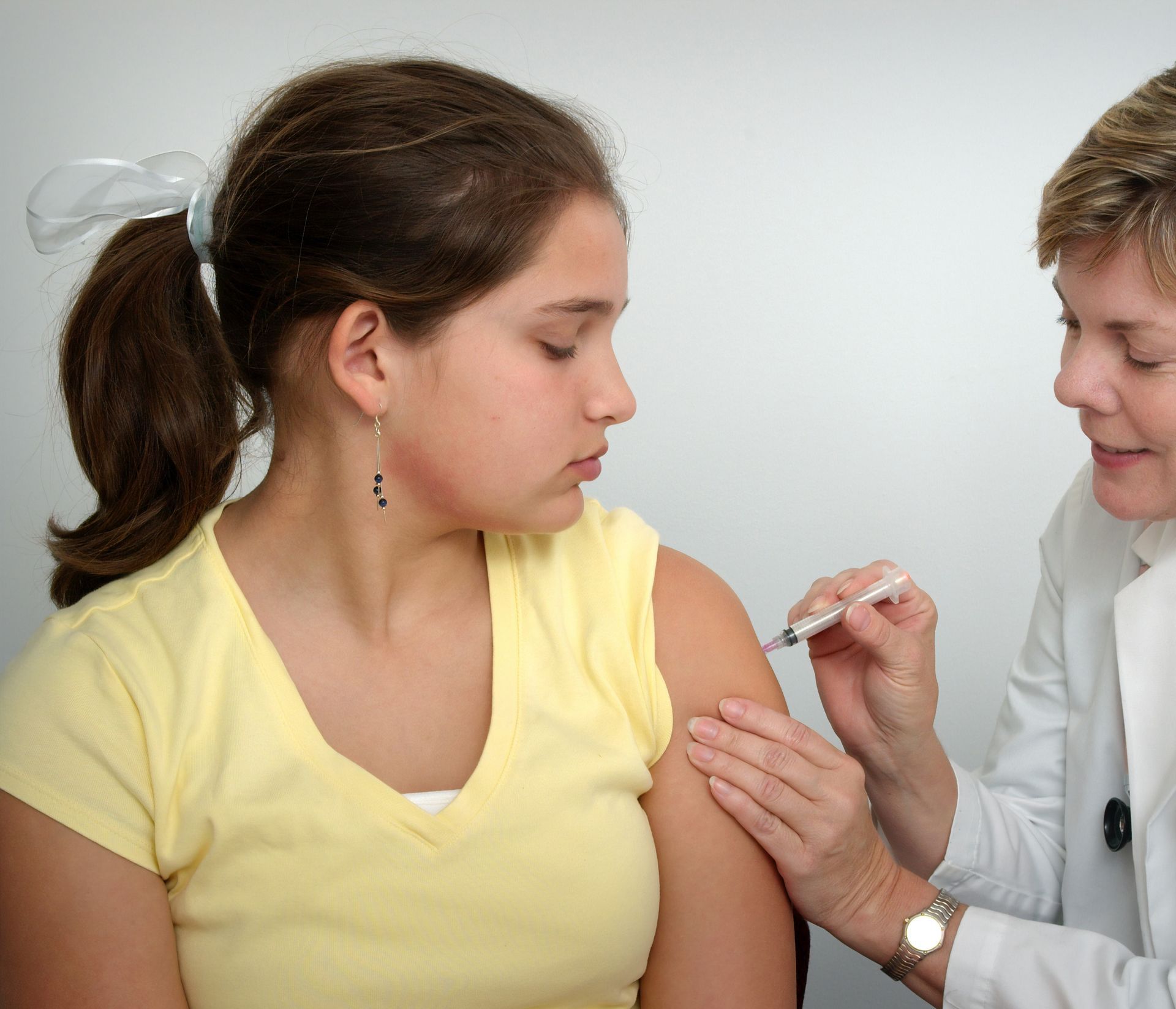 Woman giving a young person a shot in the arm. The young person looks away. White background.