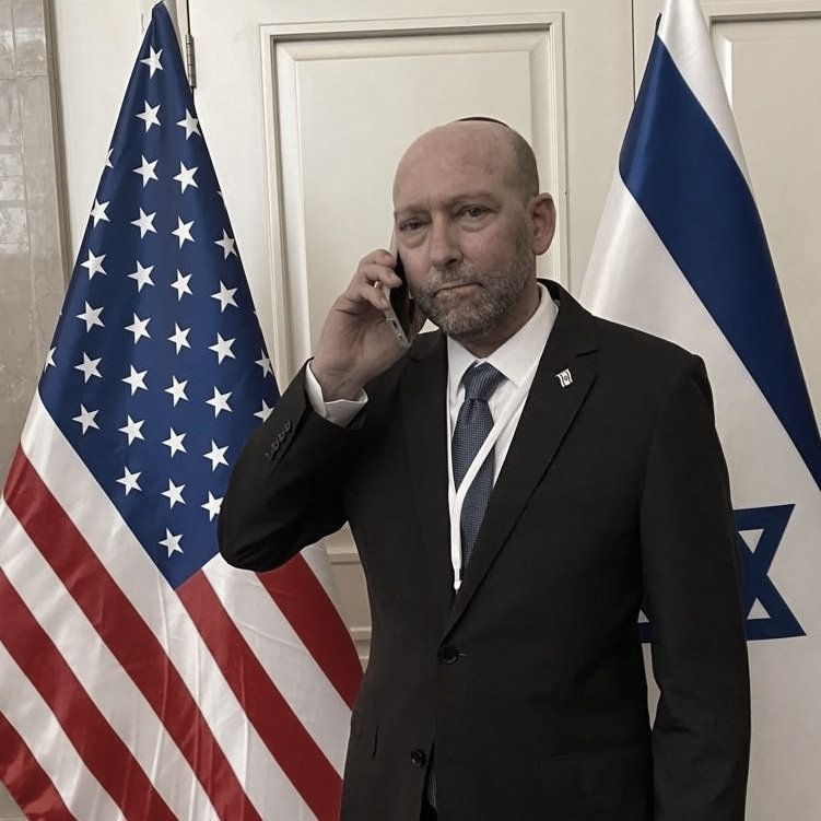 גבר בחליפה ועניבה מדבר בטלפון סלולרי מול שני דגלים
