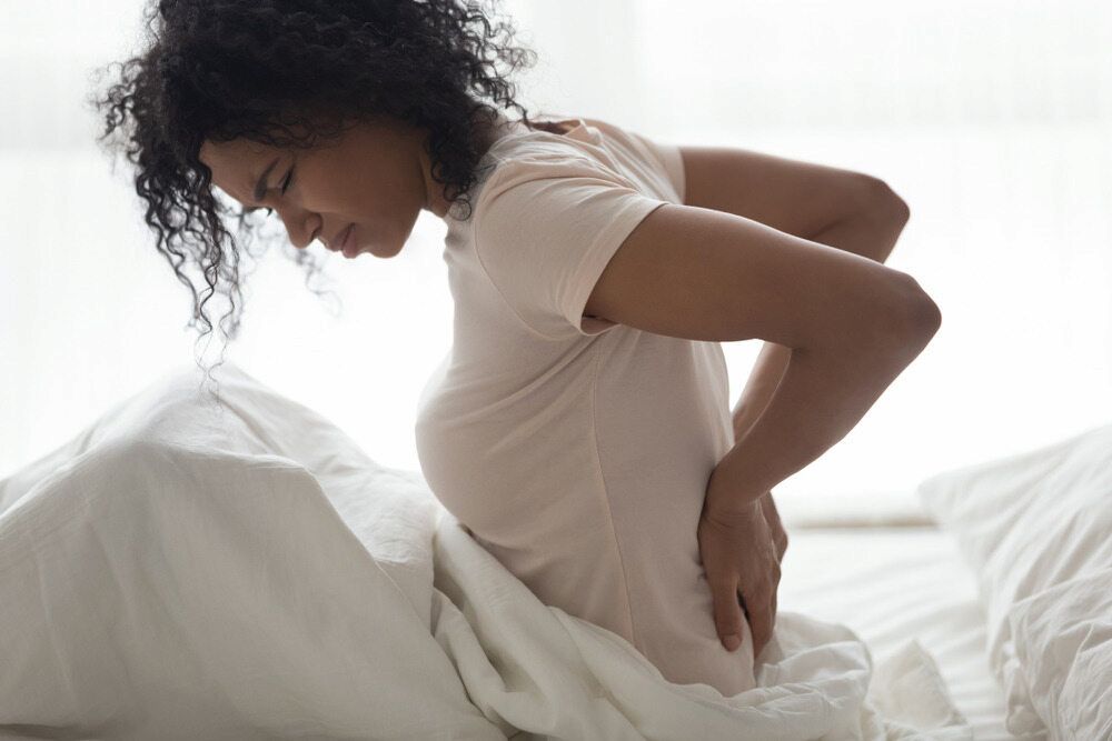 A Woman is Sitting on a Bed Holding Her Back in Pain — Life24 Health Bowen Therapy In Lavington, NSW