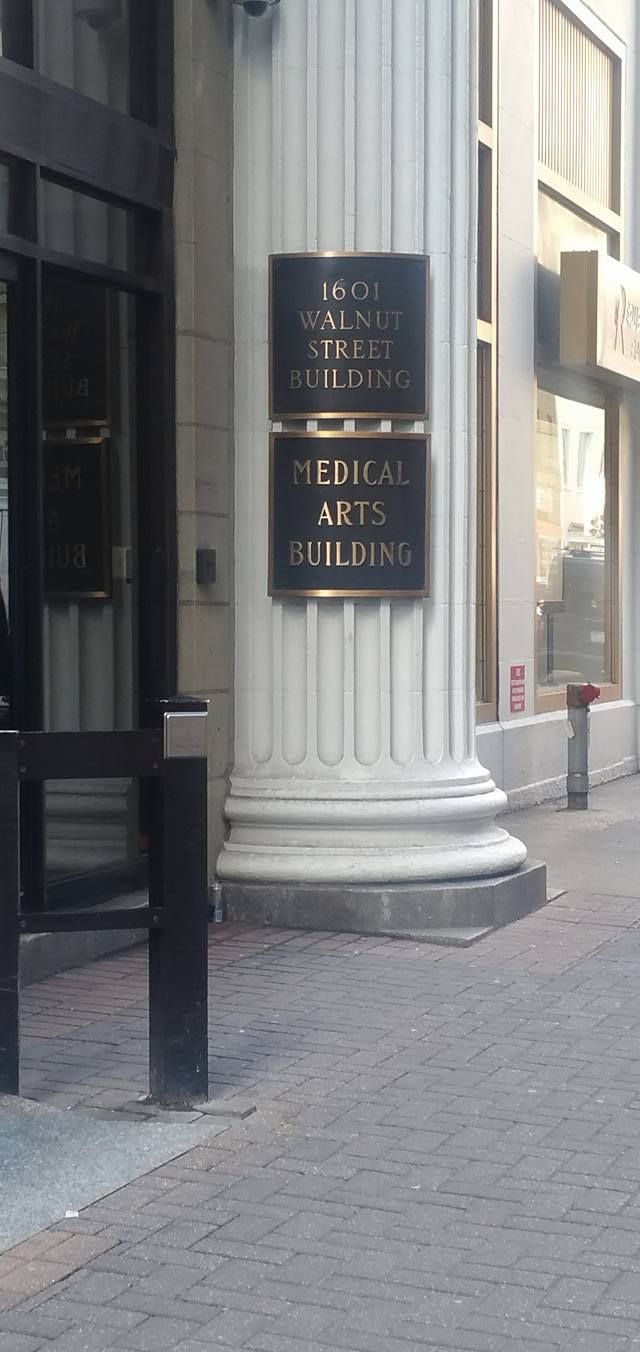 Black sign with gold lettering on a white column beside a building entrance on a cobblestone street.
