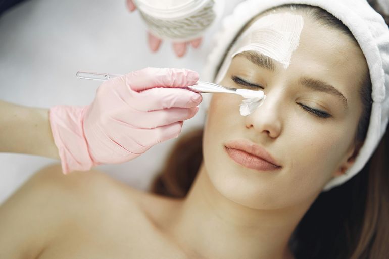 Beautician applying a facial treatment to a woman lying with eyes closed and a towel wrap