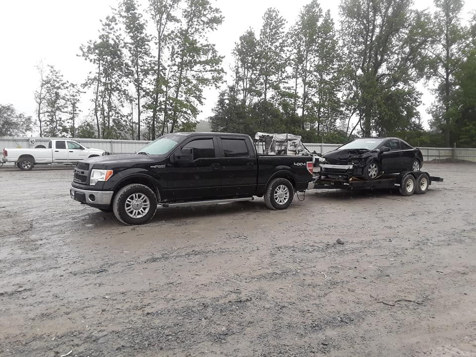 A black truck is towing a black car on a trailer in a parking lot.