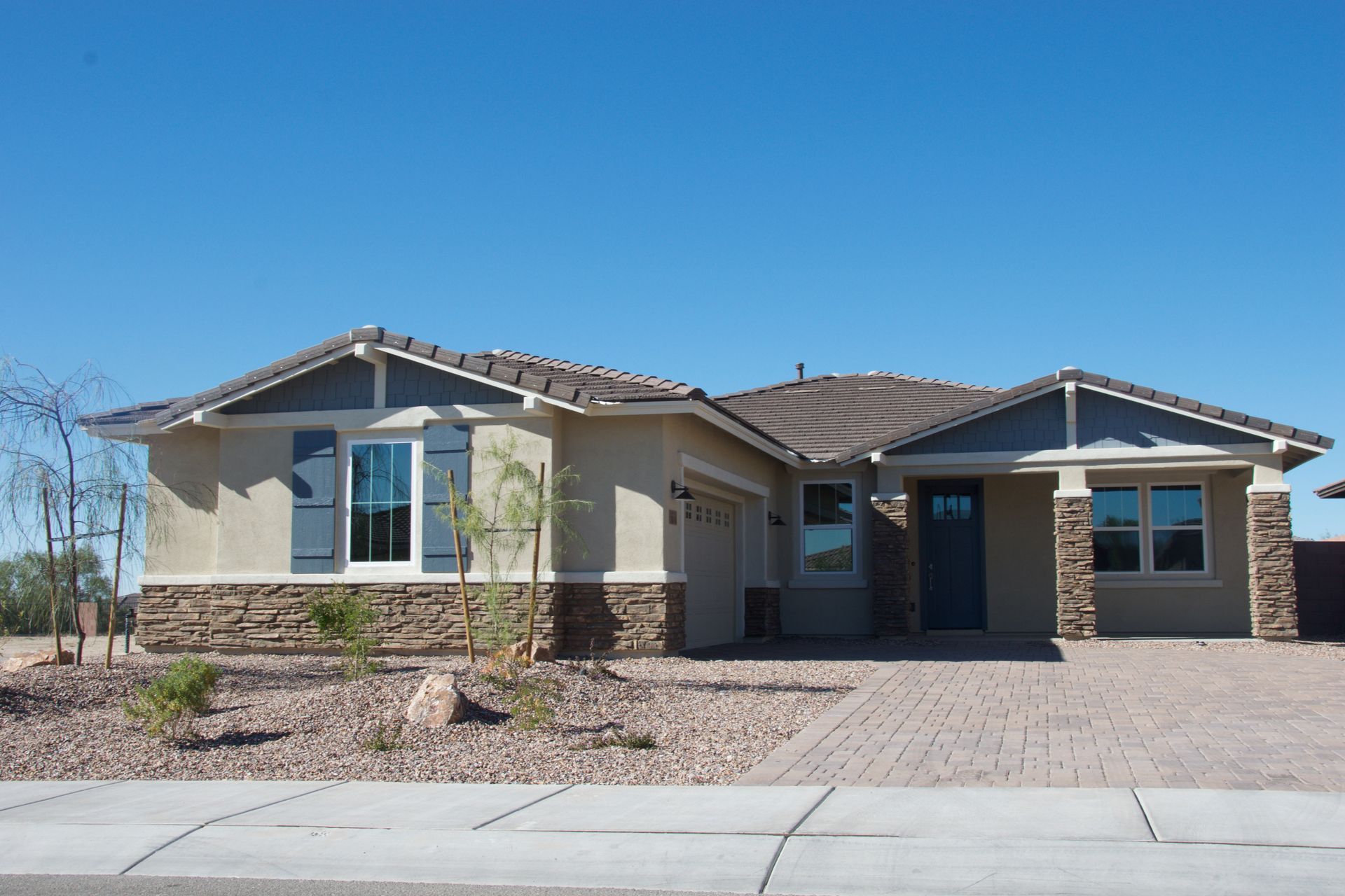 Single-story tan house with stone accents, a brick driveway, and blue sky. Single-story tan house with stone accents, a brick driveway, and blue sky.