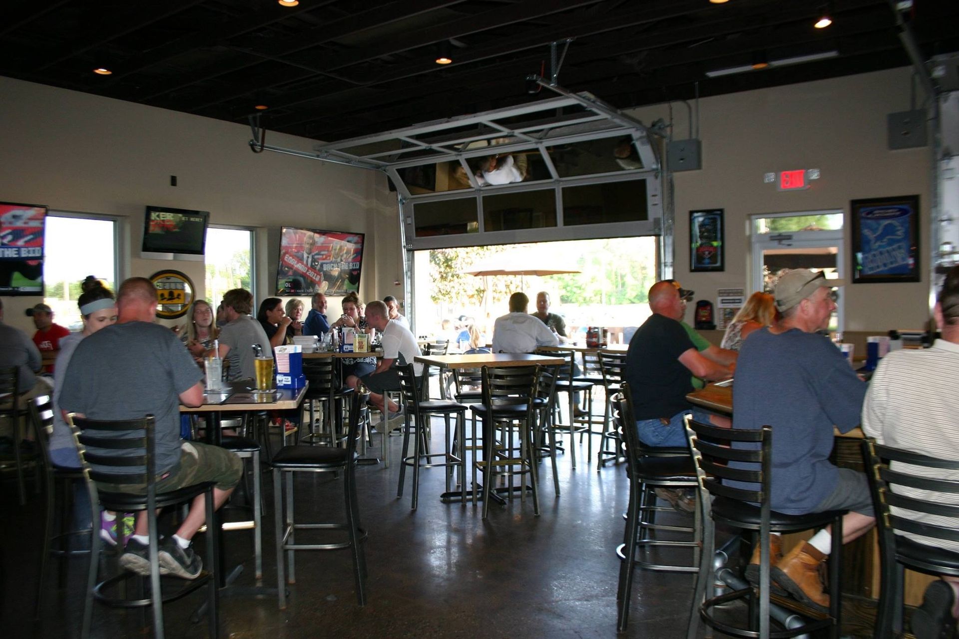 Patrons seated at tables inside a casual bar or restaurant with an open garage-style entrance