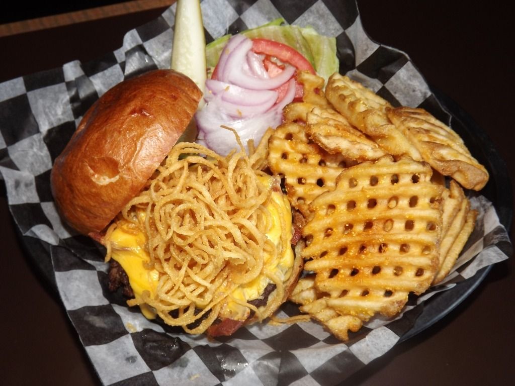 Burger with melted cheese, waffle fries, onion rings, and a pickle on checkered paper tray