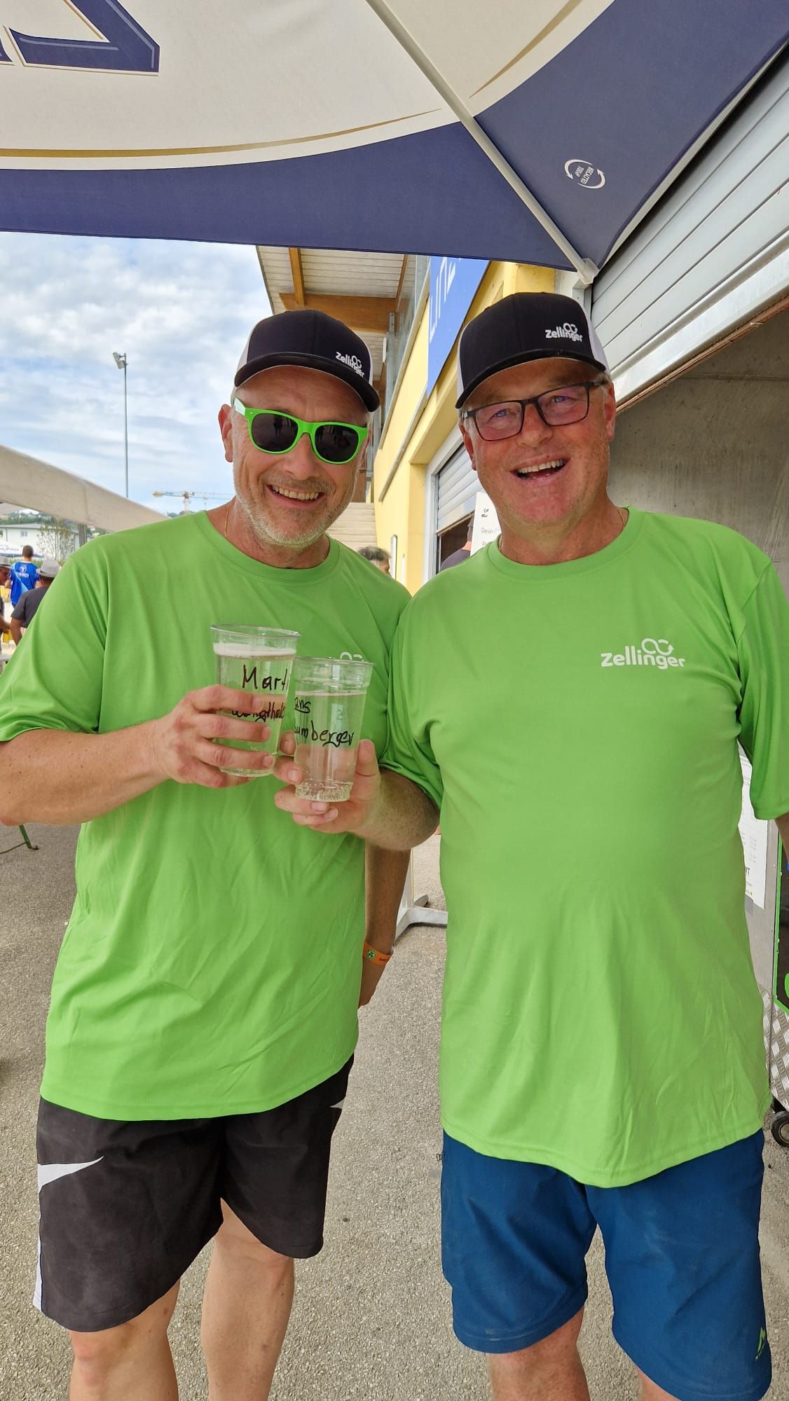 Two men in green shirts and hats, smiling, holding drinks outdoors.