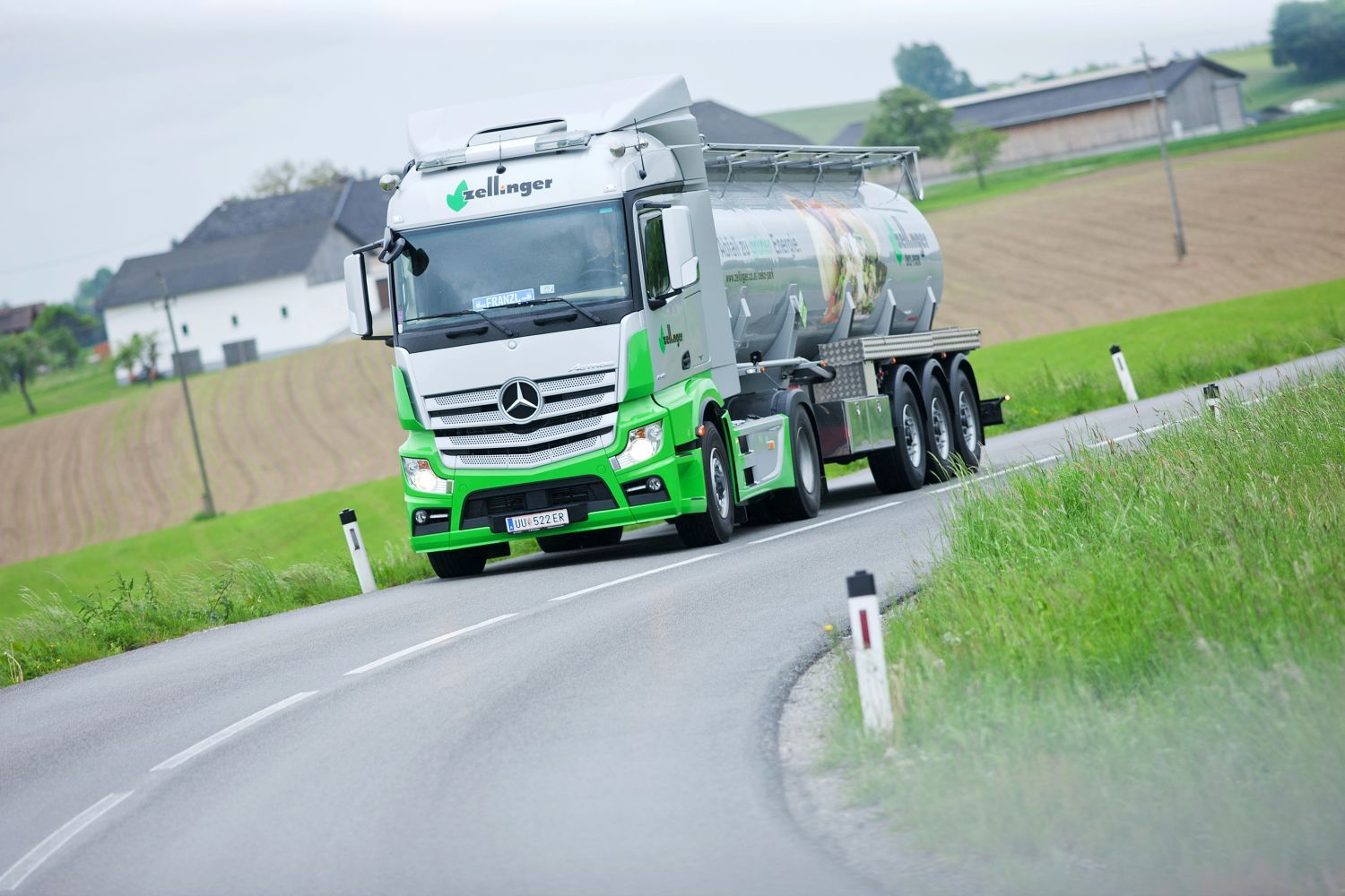 Mercedes tanker truck on a winding road, green and silver with farm buildings in background.