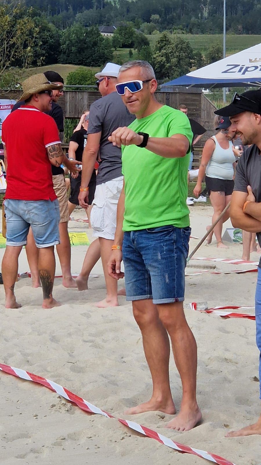 Man in green shirt and denim shorts on a sandy beach, checking watch, with others in background.