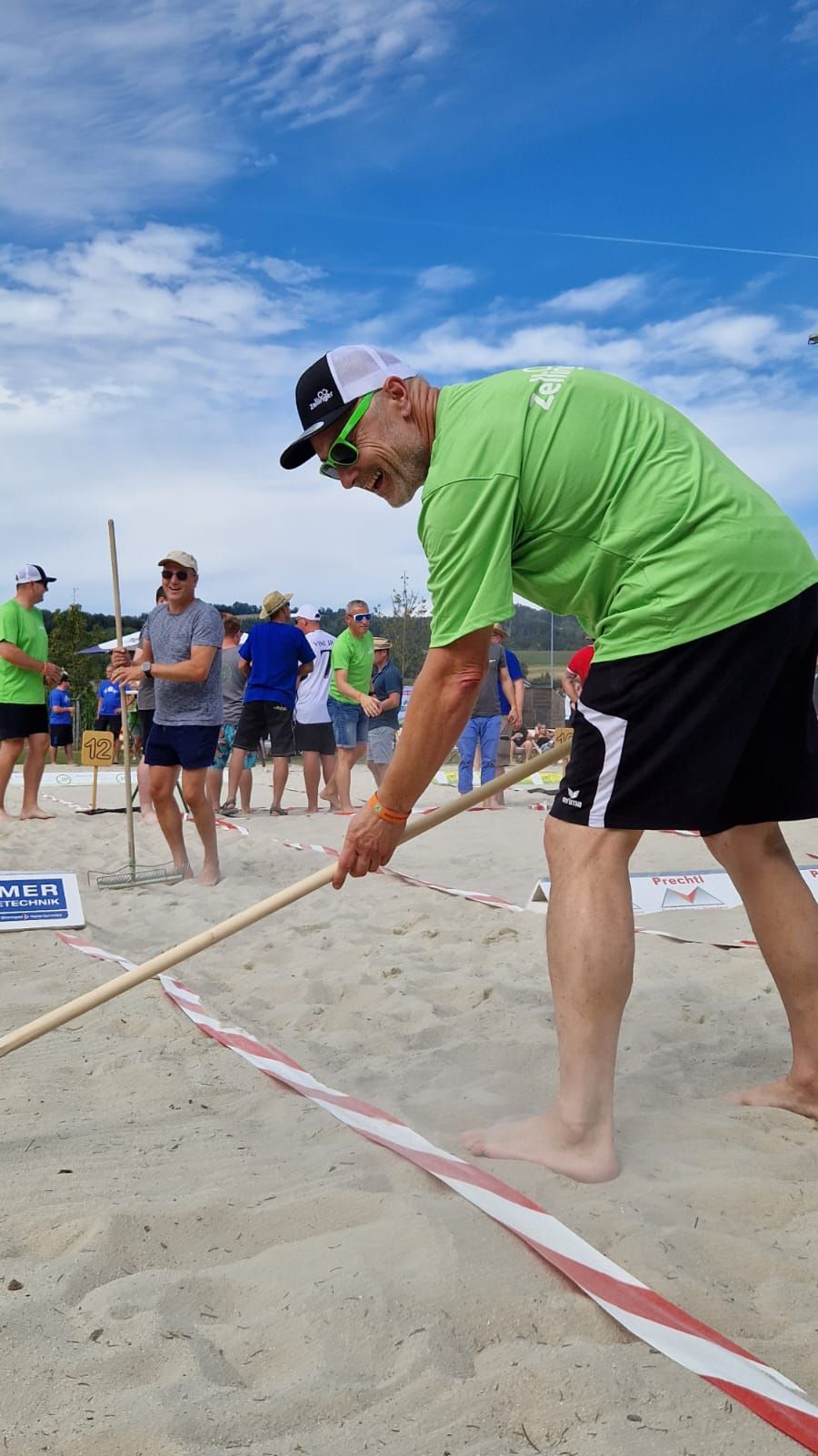 Man in green shirt and sunglasses on a beach marking a line in the sand with a stick; other people watch.