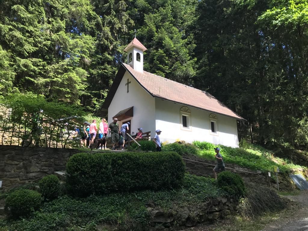 Small white church with a cross, people standing outside, set in a forest, sunny day.