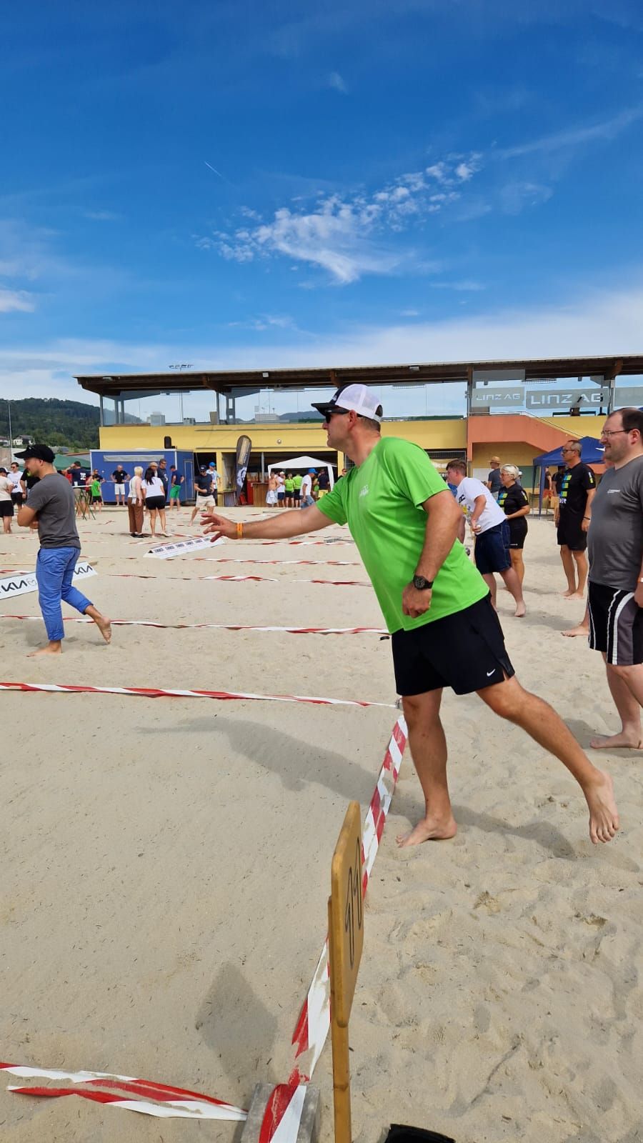 Man in green shirt throws dart on sandy beach; other players nearby. Sunny day.