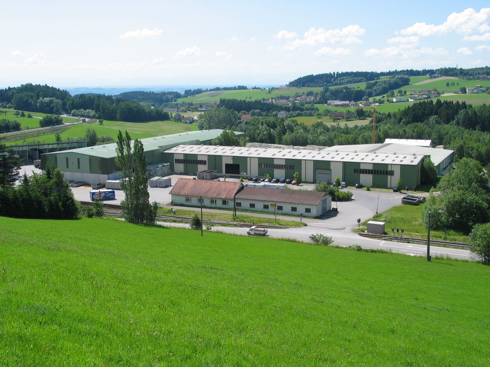 Factory buildings in a green field, rolling hills in the background.