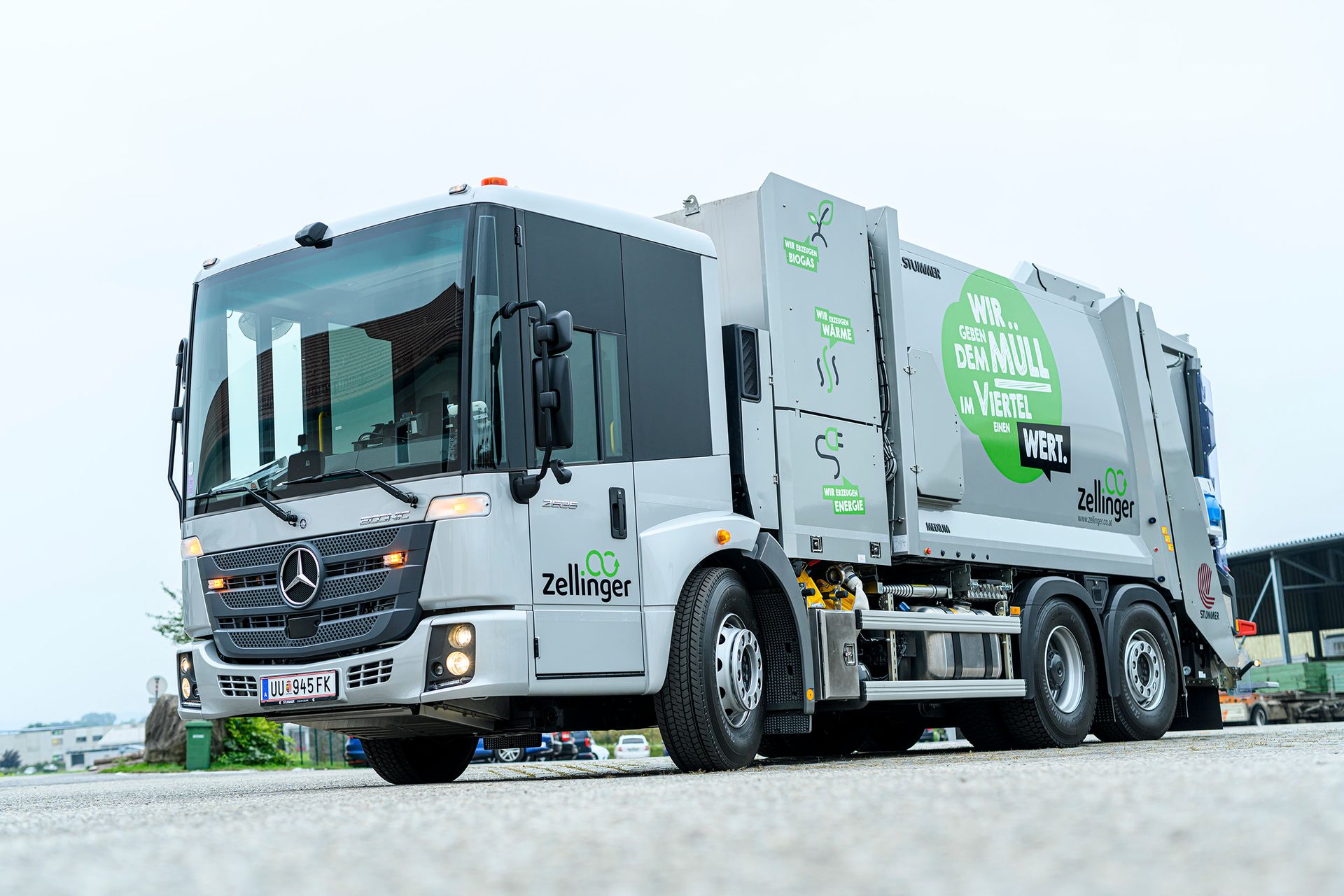 Silver Mercedes garbage truck, on a paved road, with green accents, clear sky.