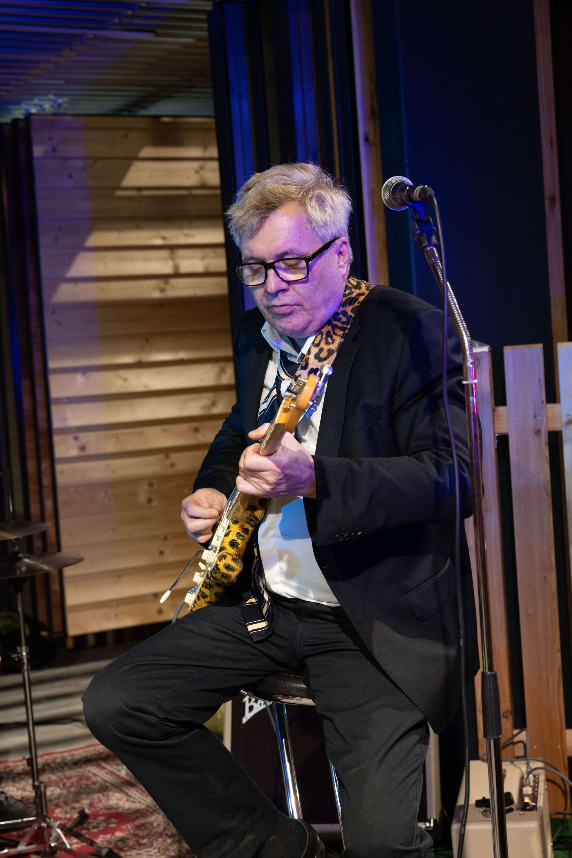 Mann mit Brille und dunklem Blazer spielt eine Leoparden-Print Gitarre. 