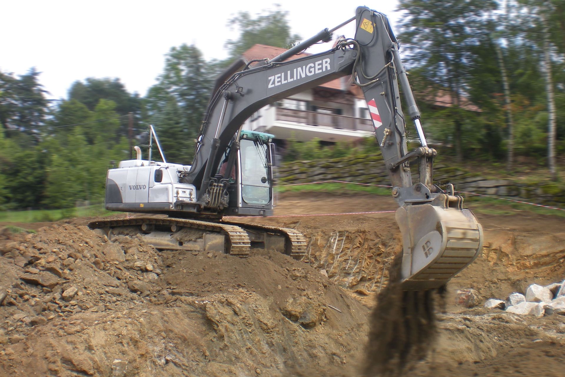 Excavator digging earth on a construction site with a house in the background.