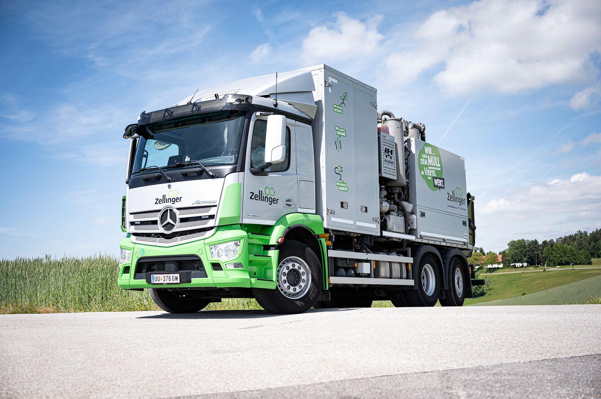 Green and silver garbage truck parked on road, with blue sky and field in background.