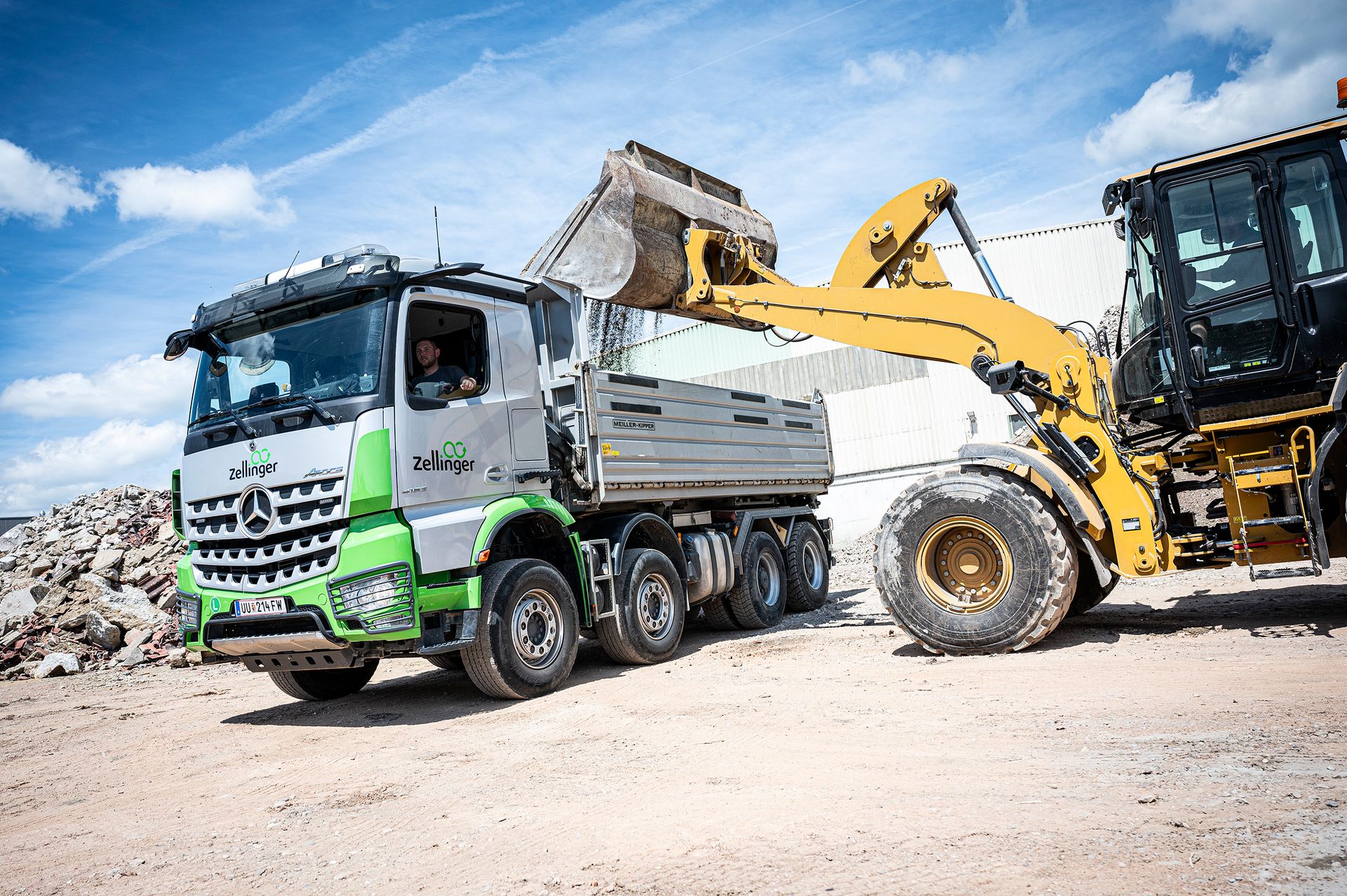 A yellow excavator loading dirt into a green and silver dump truck. Sunny outdoor setting.
