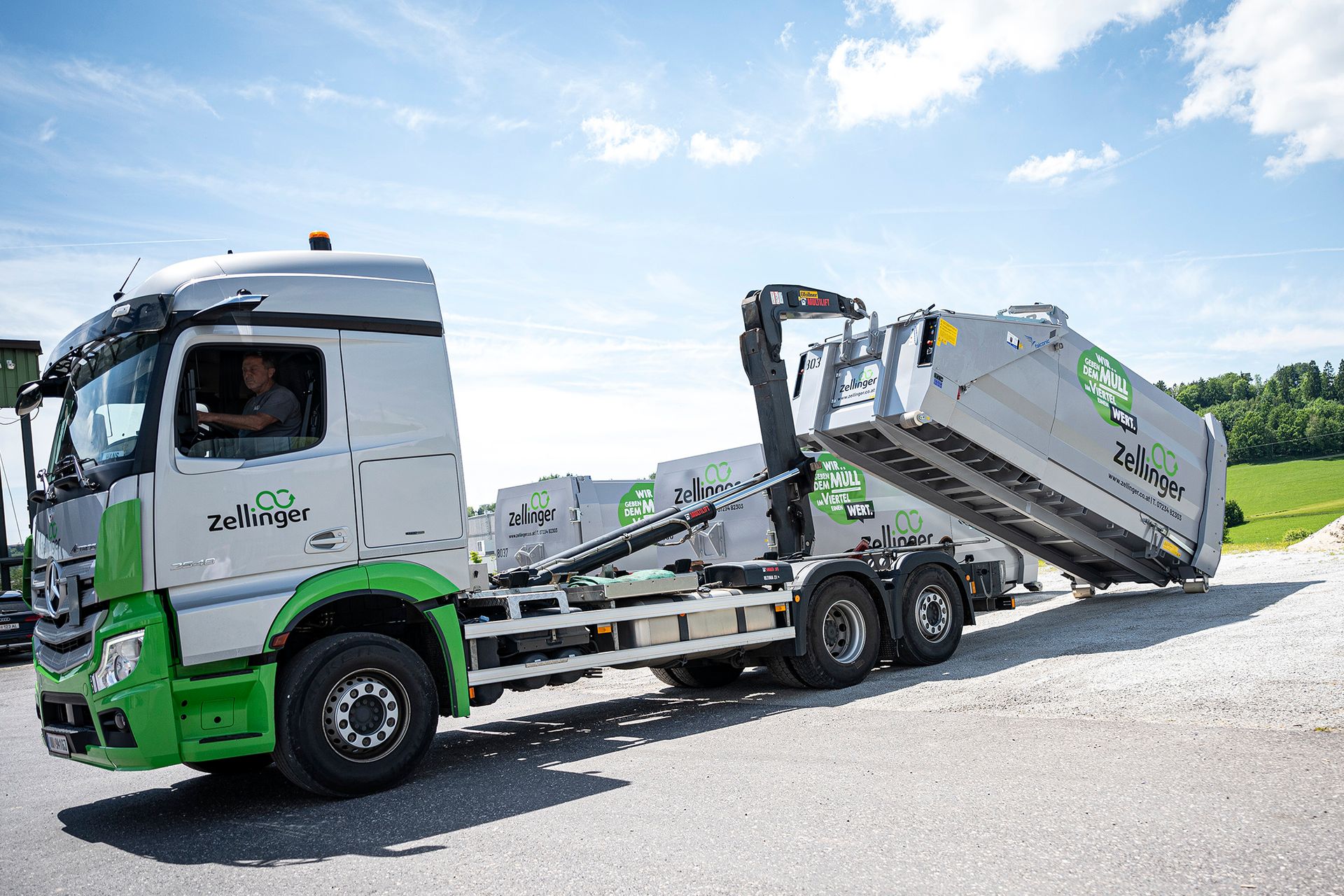 Truck with raised container, operated by a driver, for waste collection; gray and green color scheme.
