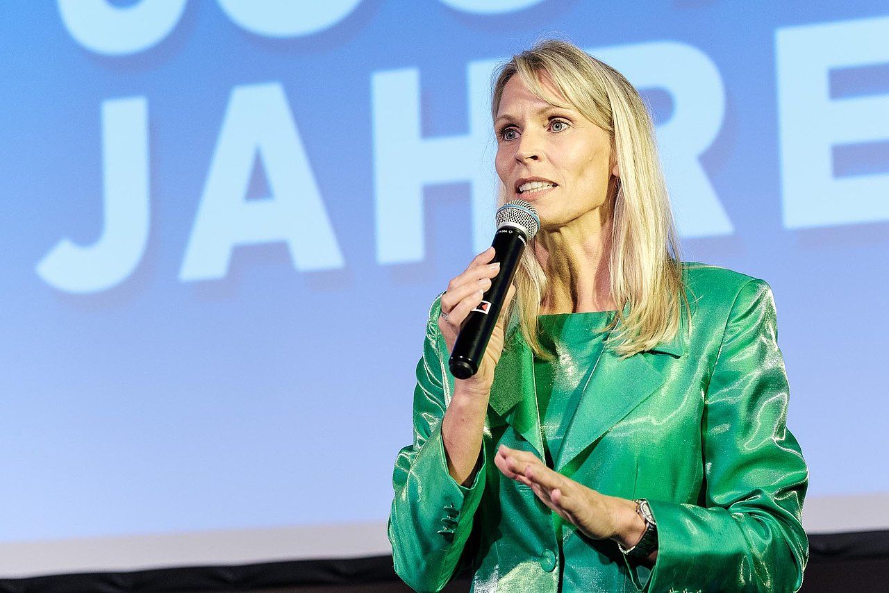 Woman with blonde hair in a green suit speaks into a microphone. Blue backdrop with 