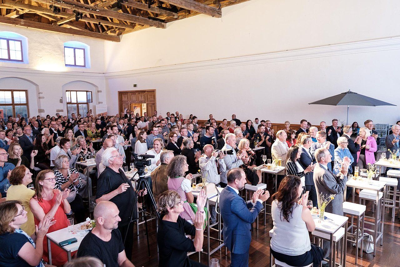 Large crowd applauding at event in spacious, well-lit hall.