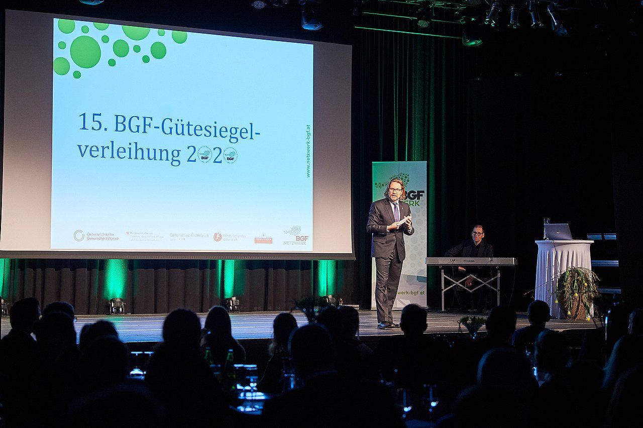 Man speaking at event, stage with screen, audience in foreground.