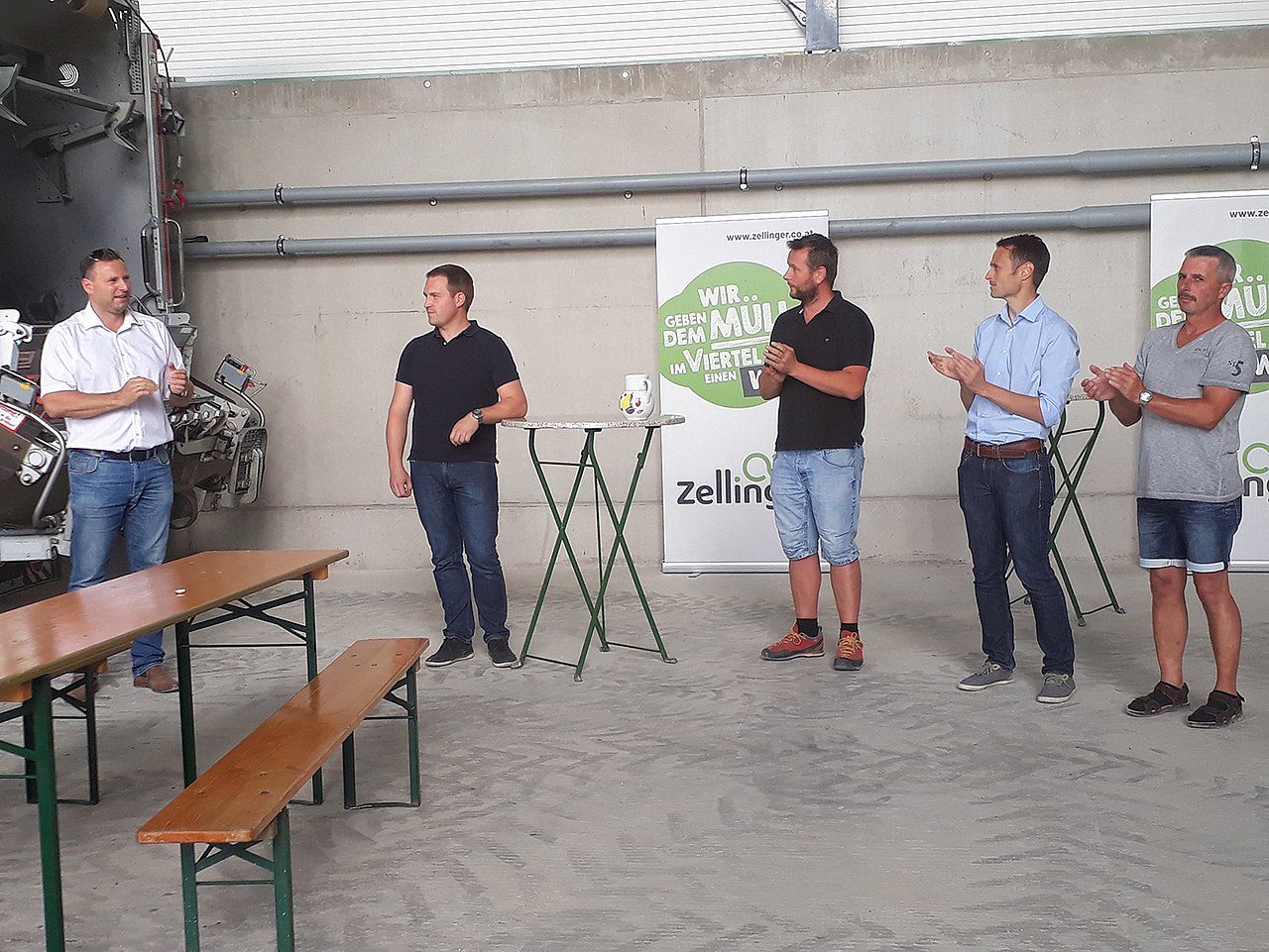 Men in casual attire at an event, clapping in front of a banner and tables inside a large structure.