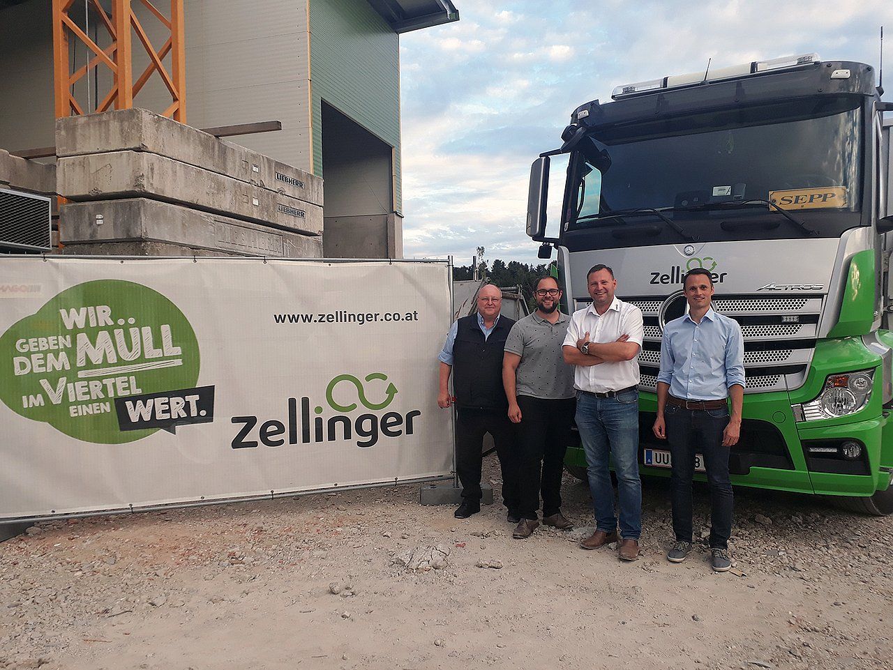 Four men stand in front of a truck and sign. Green truck, banner with logo