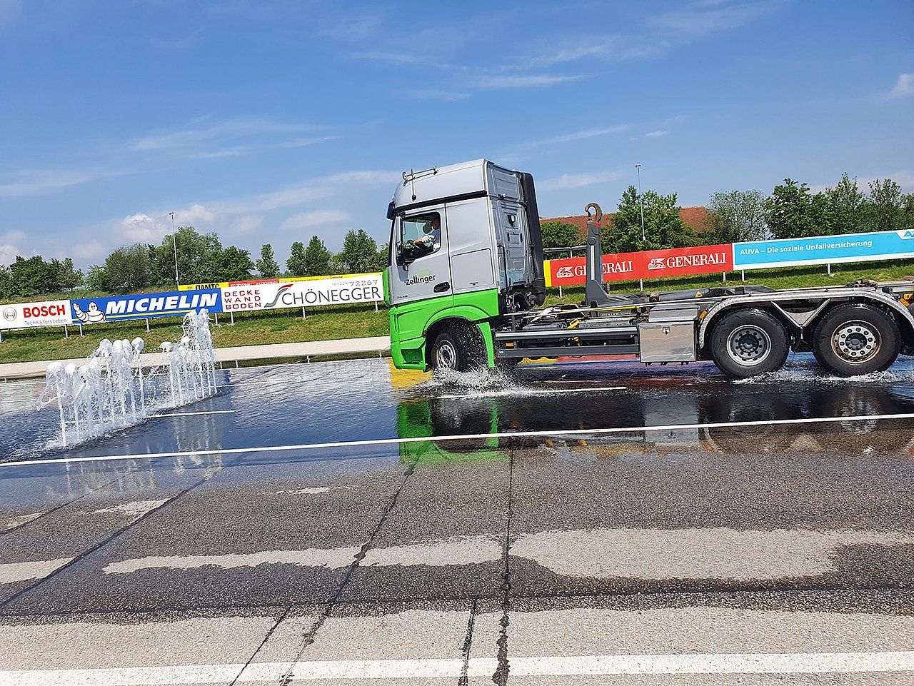 Semi-truck being washed with water, on a paved surface. Bright daylight outdoors.