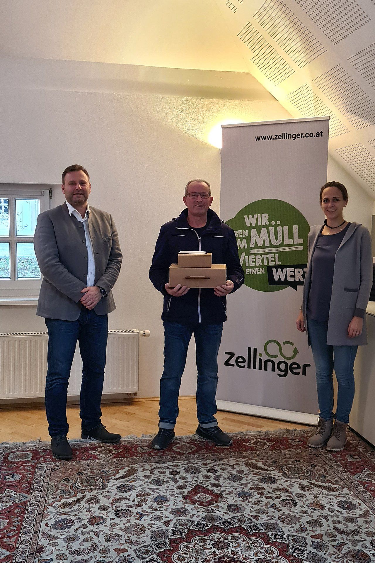 Three people standing in a room, one holding boxes, Zellinger banner in background.